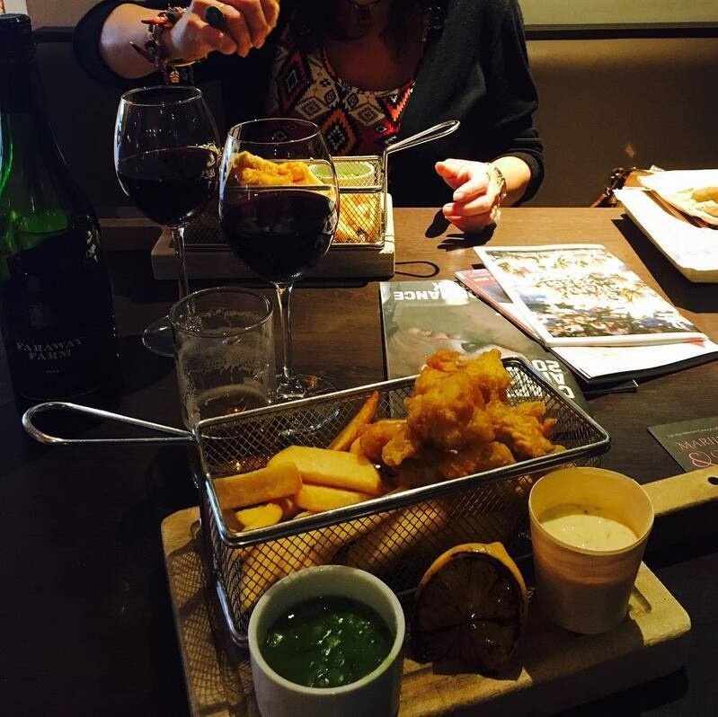Two people are enjoying fried food served in metal baskets along with two glasses of red wine and dipping sauces on a wooden table.