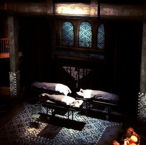 Three bodies wrapped in white cloth lie on separate metal tables in a dimly lit room with patterned tile flooring and stained glass windows.