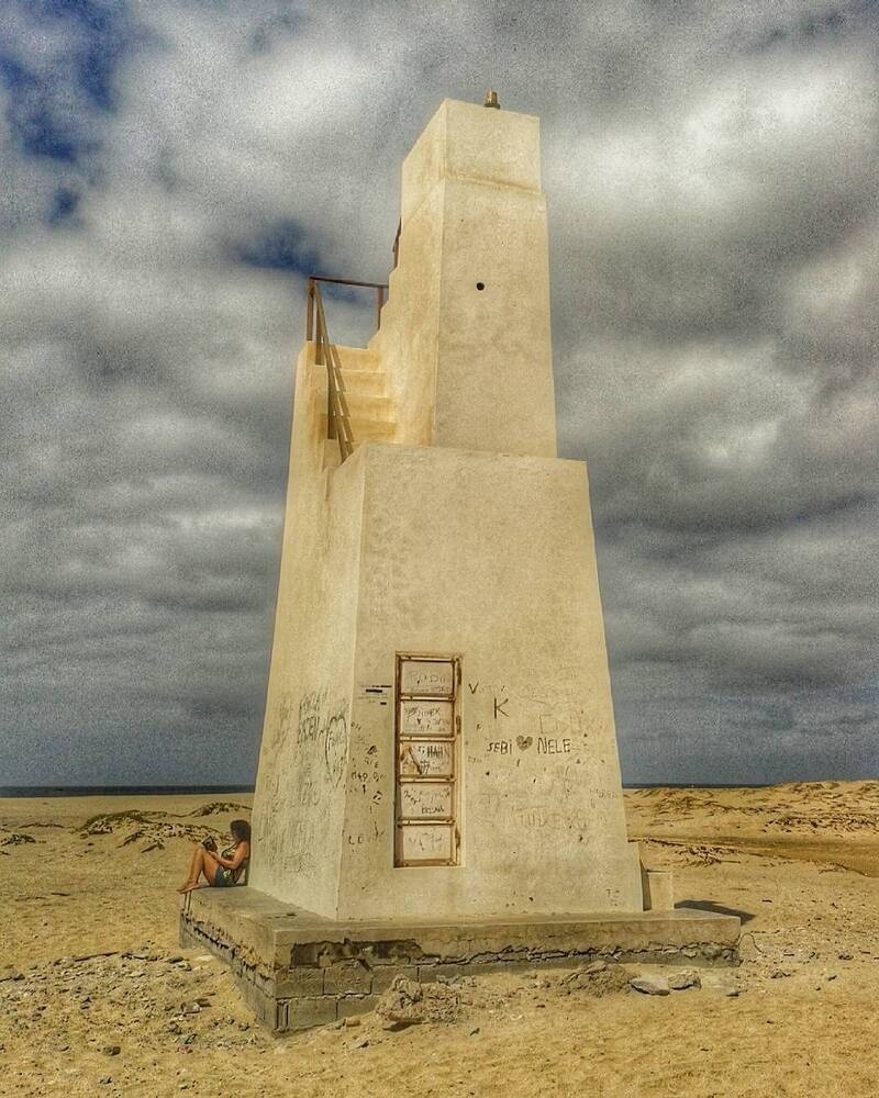A tall, white, weathered concrete structure with stairs and graffiti stands alone on a sandy beach under a cloudy sky.
