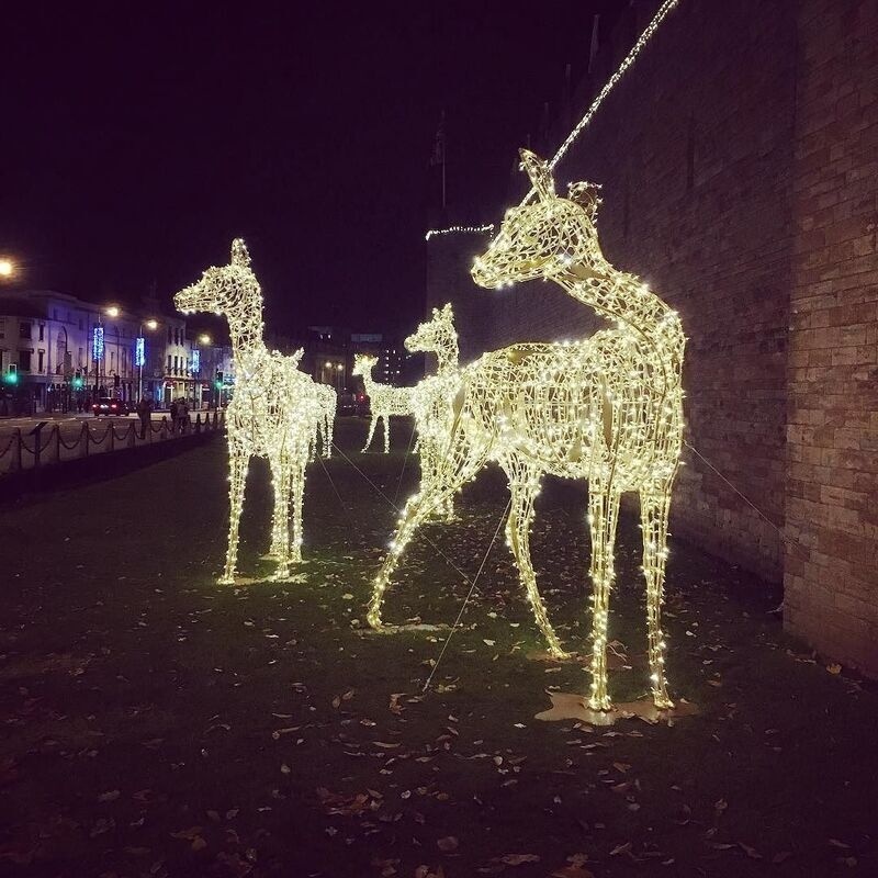 Three illuminated reindeer decorations are standing on a grassy area at night near a brick wall and a street with buildings and lights.