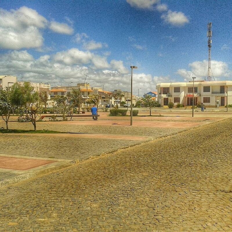A spacious, cobblestone-paved urban park area with benches, small trees, and surrounding modern buildings is shown under a partly cloudy sky.
