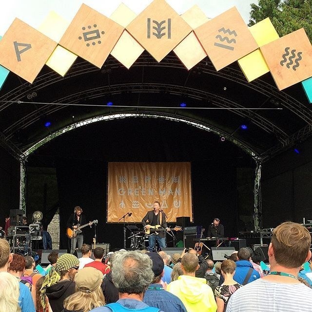 A live band is performing on an outdoor stage decorated with geometric symbols, while a crowd watches.