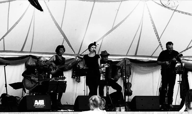 A band of musicians is performing live on stage, with various instruments including a guitar, accordion, and violin.
