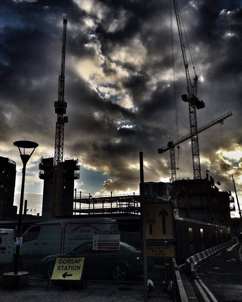 Dark clouds loom over a construction site with cranes towering above and signs pointing towards Gorsaf Station.