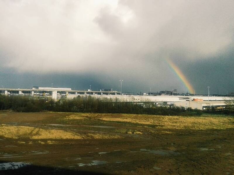 A rainbow appears over an industrial area with a cloudy sky and a mix of grassy and muddy land in the foreground.