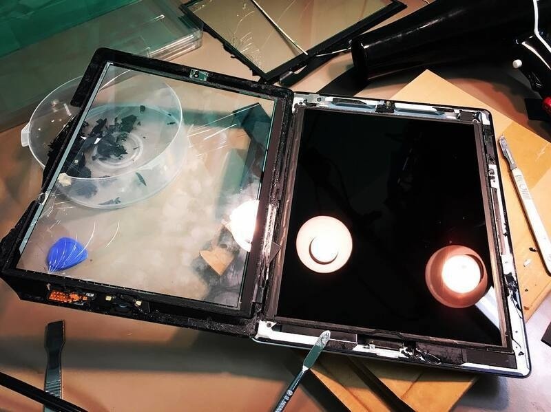 A disassembled tablet or laptop screen is shown on a workbench surrounded by tools and components.