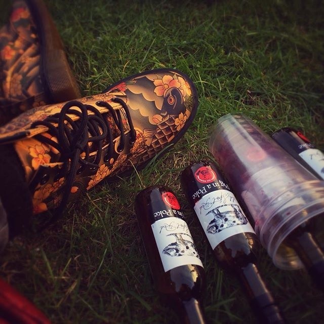 A pair of intricately patterned boots rest on grass next to three small bottles of what appears to be wine and a stack of plastic cups.