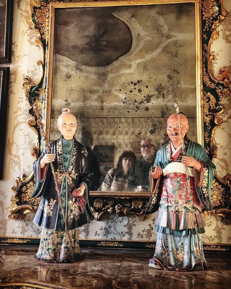 Two intricately dressed porcelain figurines stand in front of an ornate, gold-framed mirror that reflects two people and part of the room behind them.
