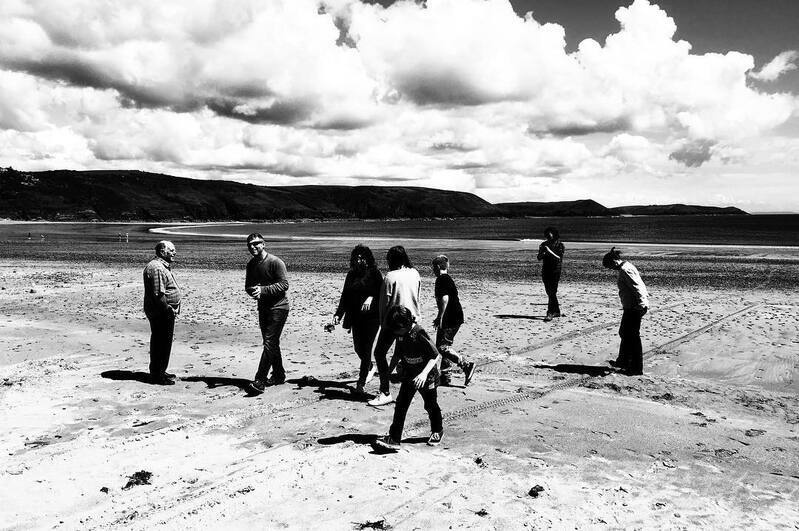 A group of people of various ages are walking and standing on a sandy beach under a cloudy sky with hills in the background.