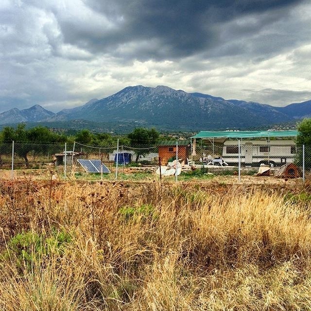 A fenced area with a small shed, solar panels, and various items sits in front of a green landscape and a mountain under a cloudy sky.