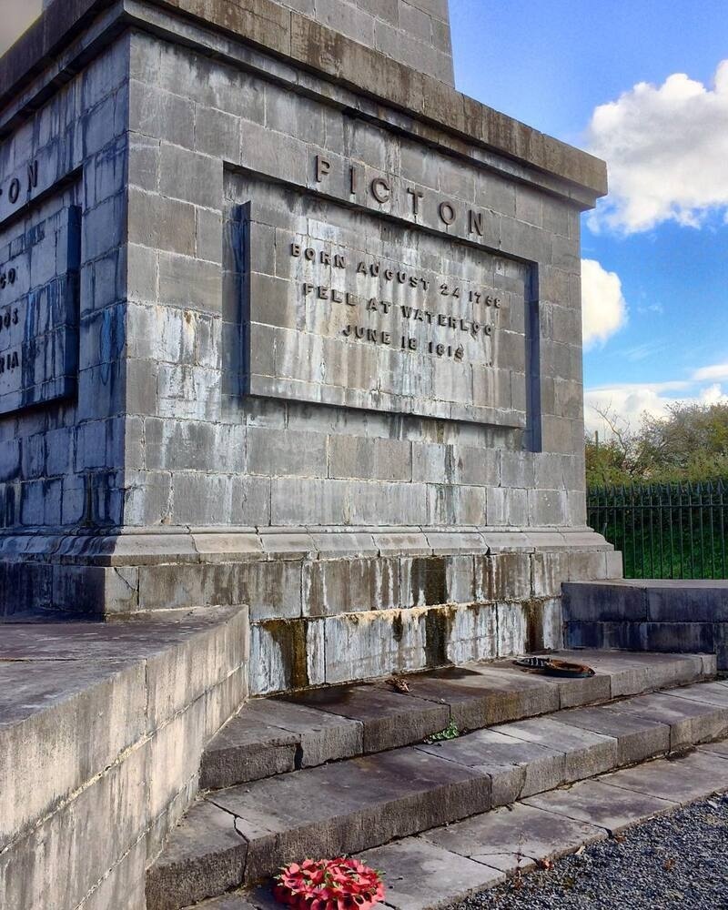 A stone monument engraved with the name Picton and details about birth and death dates stands on a paved area with a few fallen red flowers in front.