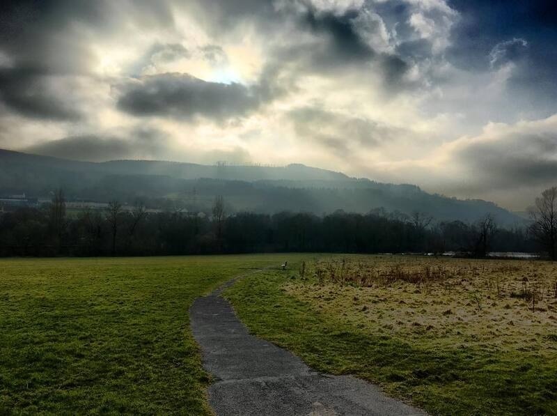 A winding path leads through a grassy field toward a misty, tree-covered hillside under a dramatic, cloudy sky.