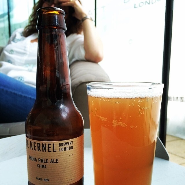 A partially filled glass of amber-colored beer is placed next to a bottle of Kernel Brewery's India Pale Ale with a person relaxing in the background.