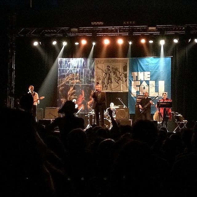 A band is performing live on stage with multiple musicians playing instruments under bright stage lights and banners displaying THE FALL in the background.