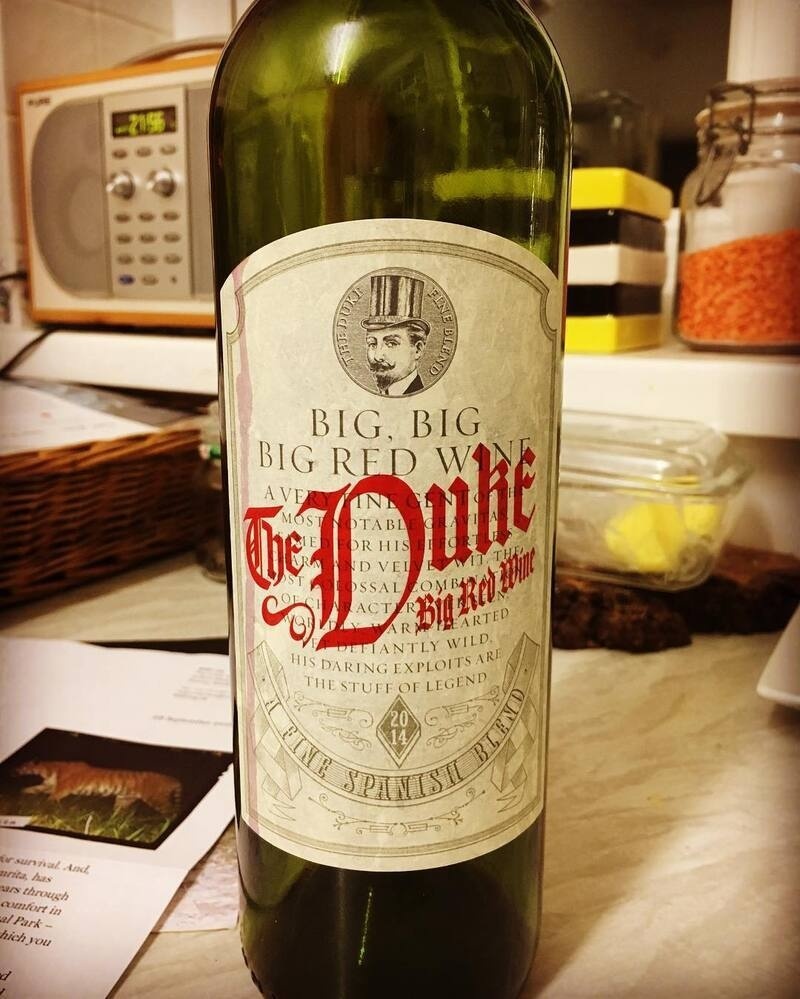 A wine bottle labeled The Duke Big Red Wine with a vintage year of 2014 is placed on a kitchen counter near a microwave and some containers.