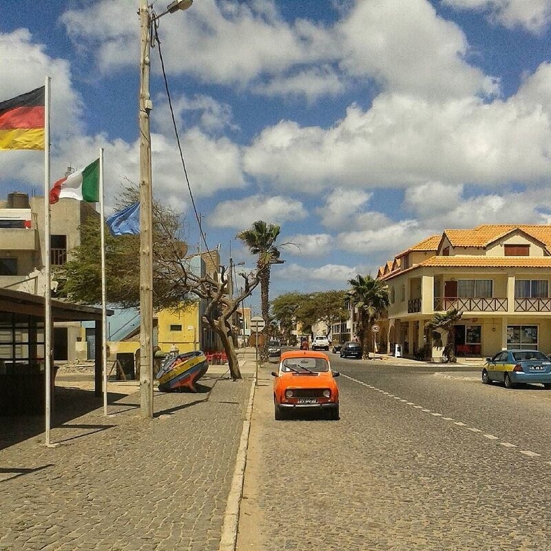 A street scene features an orange vintage car parked on a cobblestone road lined with palm trees, buildings, and several flags including those of Germany and Italy.