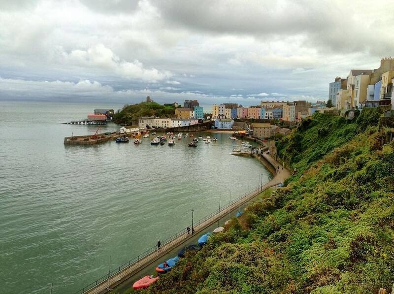 A coastal town with colorful buildings, a small harbor filled with boats, and a green hillside overlooking the sea under a cloudy sky.