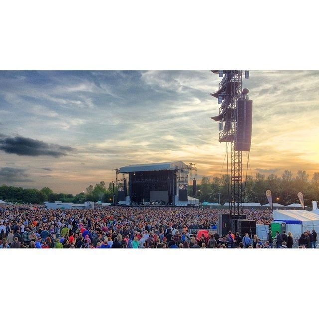 A large crowd gathers outdoors in front of a stage during a concert or event at sunset.