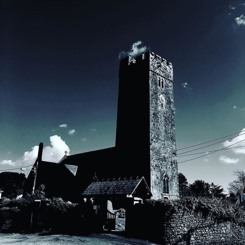 A tall, ancient stone tower with a clock stands against a dramatic sky above a dark silhouette of surrounding buildings and foliage.