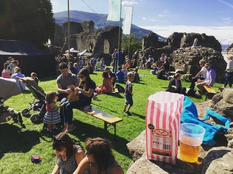 People are enjoying a sunny outdoor gathering on a grassy area near ancient stone ruins, with children playing and adults sitting on benches and the ground.