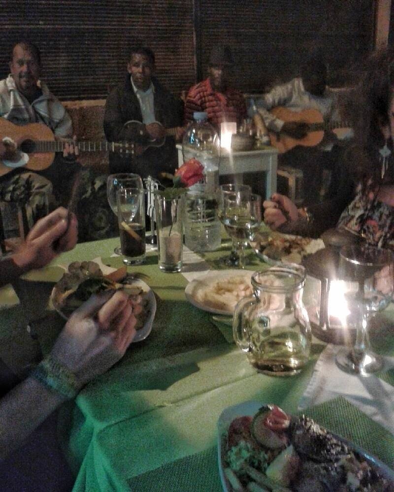 A group of people are gathered around a table eating and drinking while others in the background play guitars.