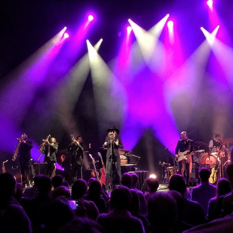 A live band performs on stage under vibrant purple and white spotlights in front of an attentive audience.