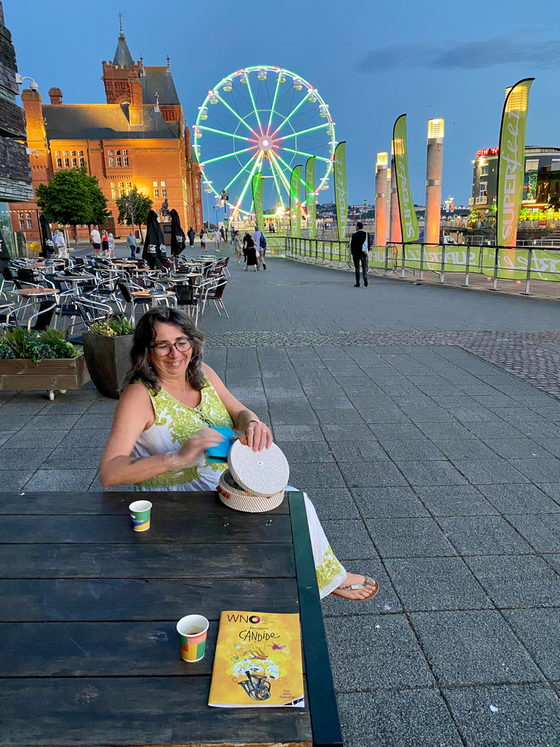 Woman sitting at table at dusk, small coffees and theatre programme on table, illuminated fairground wheel and red brick building in background
