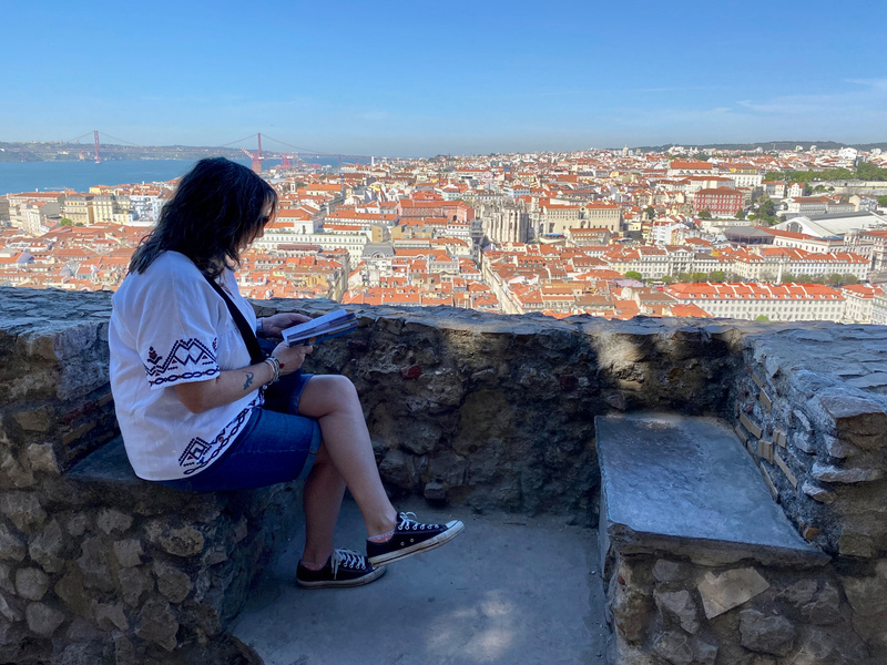 View over a city from high viewpoint, woman seated on stone wall in foreground reading a guidebook