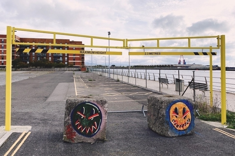 Two concrete blocks with graffiti faces blocking the entrance to an empty car park alongside open water