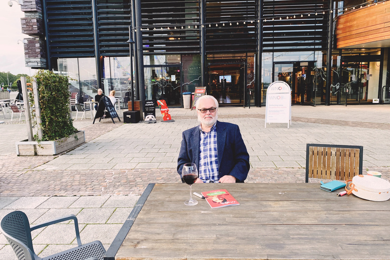 Bearded man in blue jacket and checked shirt sitting at an outdoor table with a glass of red wine