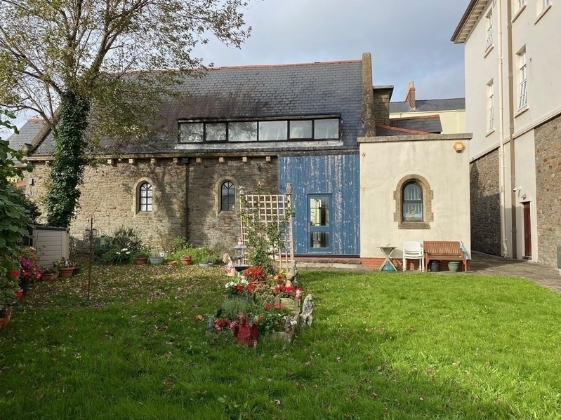 Small grassed garden behind a old stone church building. Central display of flowers, trellis and small statues