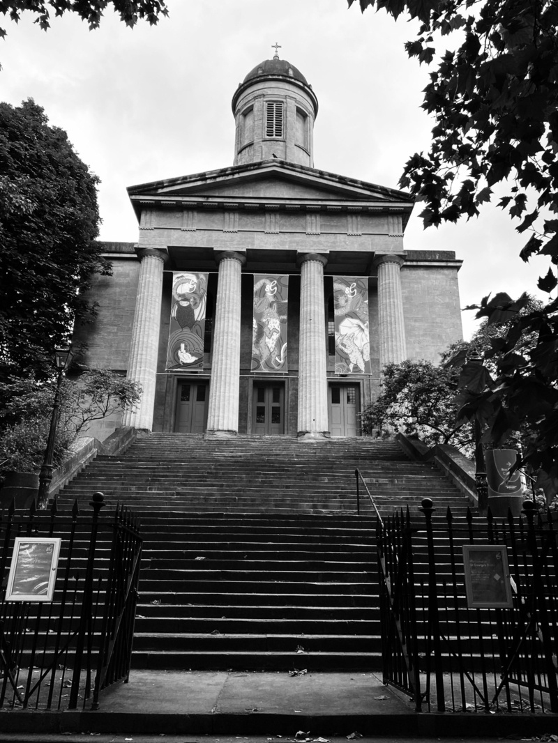 Black and white image of a neoclassical church with tall columns and a domed bell tower. The church is at the top of a long flight of steps, bordered by trees and an iron fence.