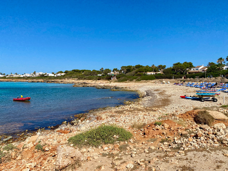 A rocky beach coastline with clear blue water, a red inflatable boat in the shallow bay, and a small cart on the beach holding kayaks and paddleboards. In the background, there are white buildings and green trees under a clear blue sky.