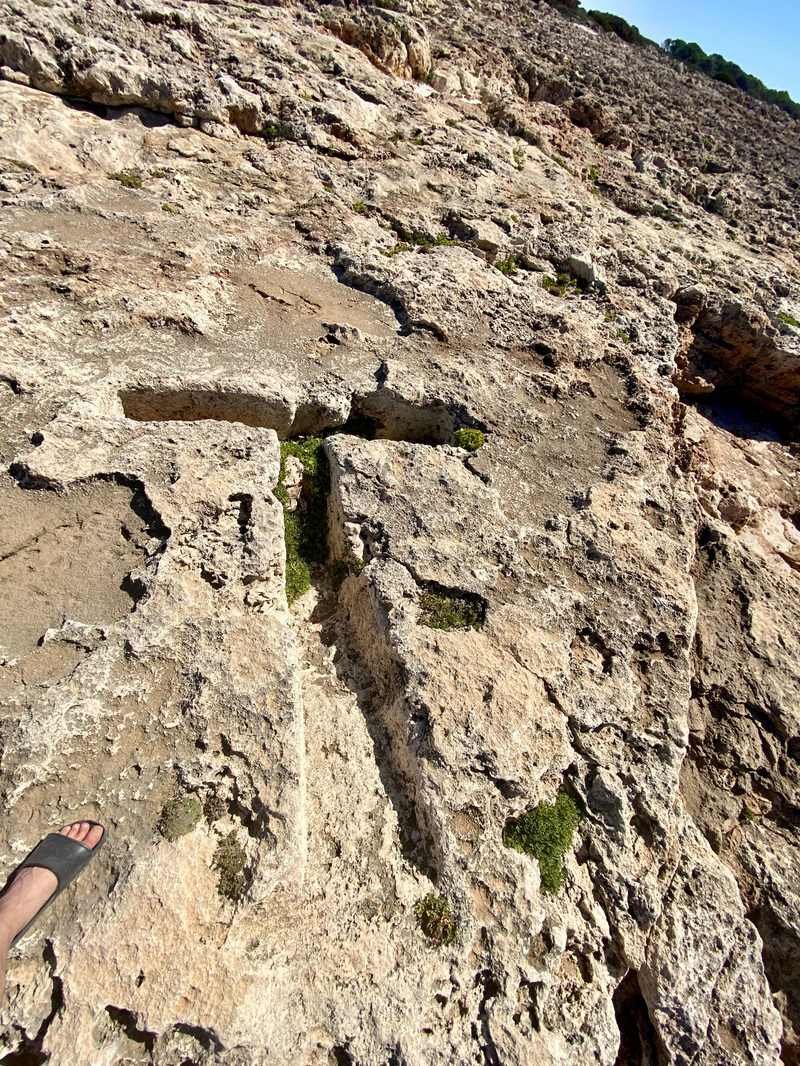 A weathered, rocky terrain with a carved T shaped deep groove. A single foot wearing a black sandal is visible at the bottom left, highlighting the uneven surface. Small patches of green plants are scattered within the rock crevices.