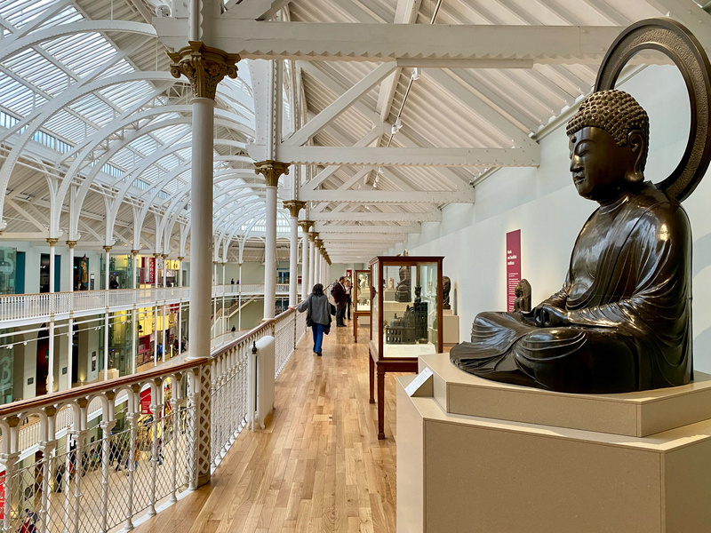 Interior of a museum gallery with a large Buddha statue, visitors walking along the balcony, display cases, and an ornate white ceiling structure.