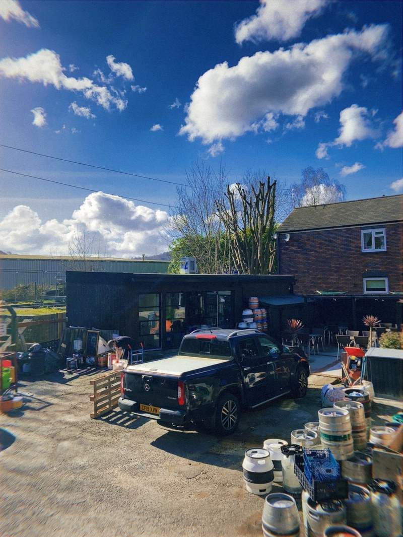 A sunny outdoor scene with a black pickup truck parked in front of a modern building with large windows and an outdoor seating area. There are multiple metal beer kegs stacked on the ground near the vehicle and around the patio. A clear blue sky with fluffy clouds