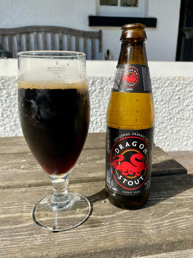 A glass of dark beer and a bottle of Dragon Stout on a wooden table. The bottle label features a red dragon and the text Original Jamaican Dragon Stout. In the background, there is a wooden bench and part of a white wall