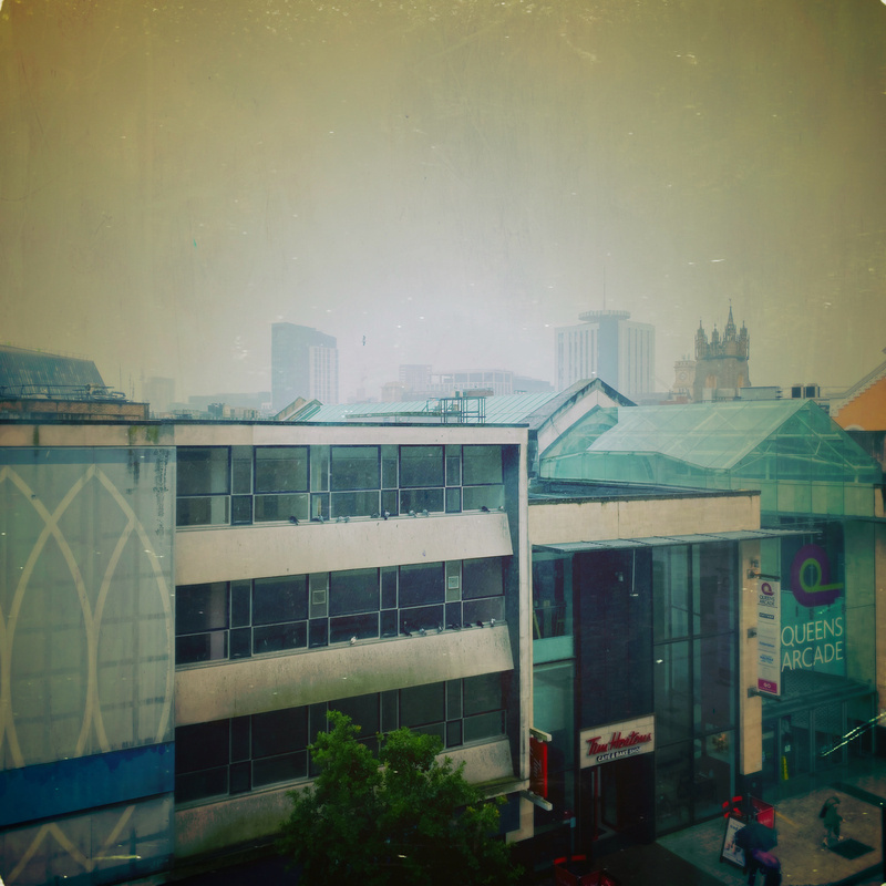 A cityscape view showing a multi-story building with a sign for Queens Arcade and a Tim Hortons cafe on the ground floor. The image features other buildings in the background, including modern high-rises and a church-like structure with spires