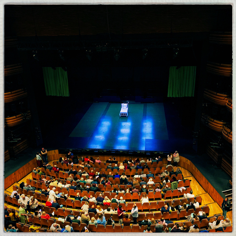 A theater view from the balcony, showing a stage with a single bed prop under blue lighting. The audience is seated and waiting for a performance to start, with a mix of empty and occupied seats.