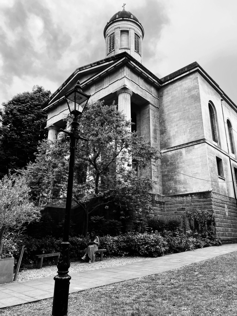 A black and white photograph of a historical church featuring prominent columns and a dome. A streetlamp stands in the foreground, and lush foliage surrounds the church. A person is seated on a bench near the pathway leading to the church. The sky is cloudy.
