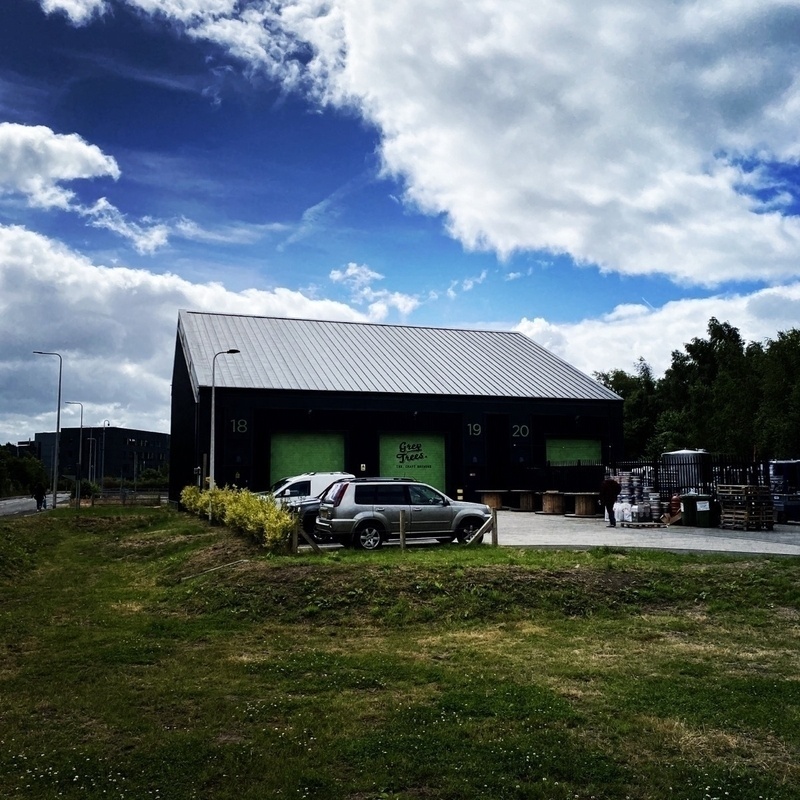 A dark-colored warehouse with the numbers 18, 19, and 20 on its doors is surrounded by parked cars and greenery under a partly cloudy sky.