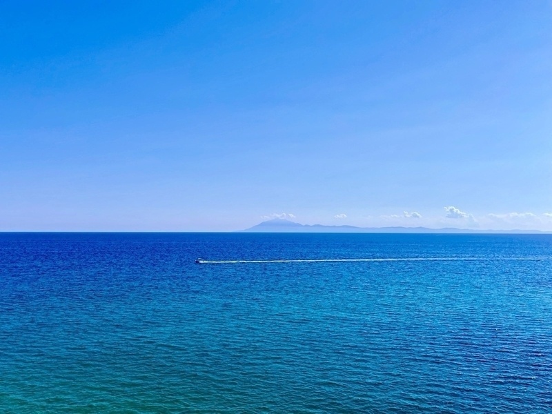 Expanse of blue sea beneath blue skies. Mountians just visible on the horizon. A small boat is causing a wake in the middle distance.