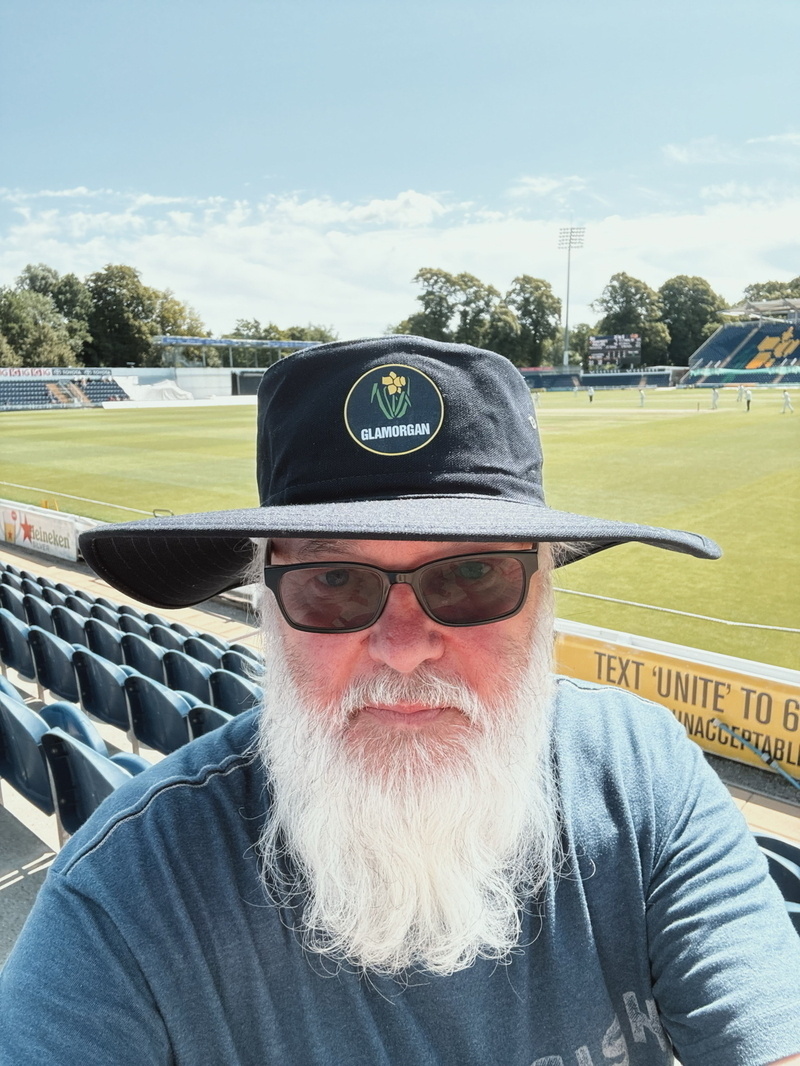 Auto-generated description: A bearded person wearing sunglasses and a large dark hat is sitting in a stadium with a cricket field in the background.
