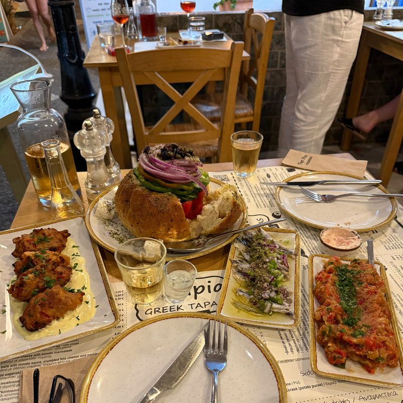 A table is set with a variety of Greek tapas dishes, including stuffed bread, grilled vegetables, and meat, accompanied by drinks.