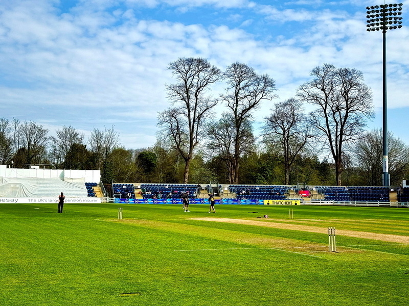 Auto-generated description: A cricket field is set up with stumps in place and a few people on the pitch, surrounded by trees and empty spectator stands.