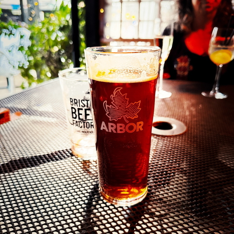 Auto-generated description: A glass of Arbor beer is on a table alongside a partially visible Bristol Beer Factory glass, with other drinks and a person in the background.