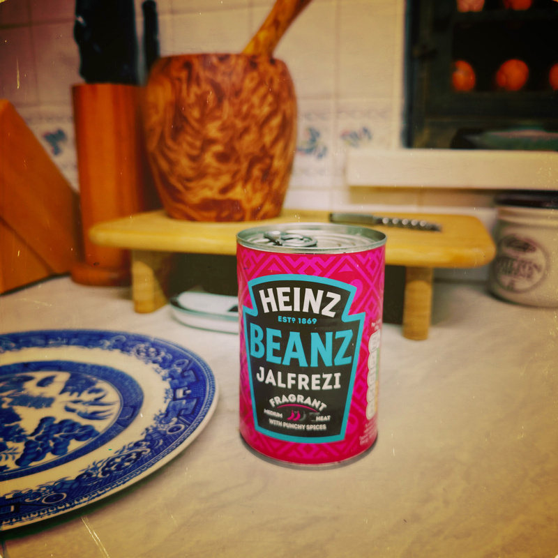 Auto-generated description: A can of Heinz Beanz Jalfrezi is placed on a kitchen countertop near a decorative plate and wooden utensils.