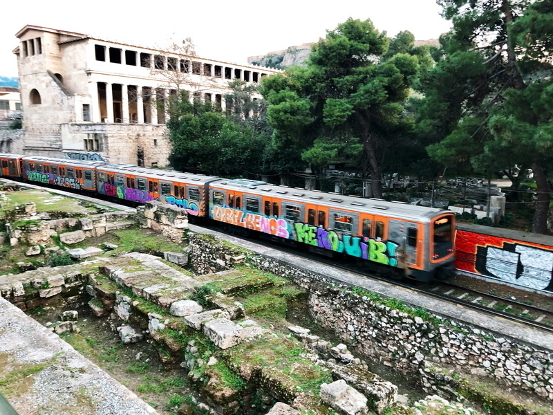 Auto-generated description: A graffiti-covered train passes through ancient ruins with trees and a historic building in the background.