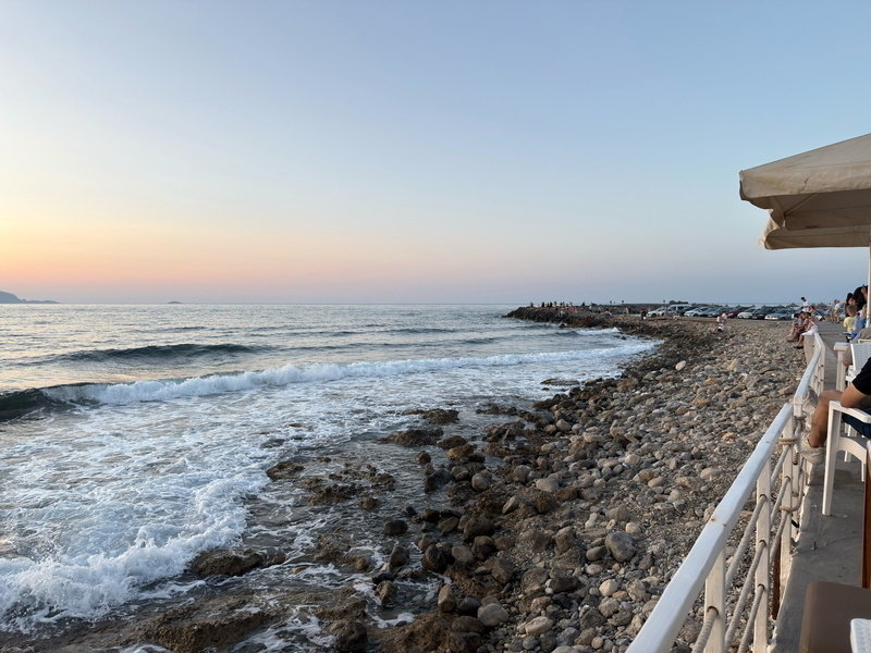 Auto-generated description: A scenic coastline with gentle waves crashing onto a rocky shore under a pastel sunset sky, viewed from a waterside terrace.