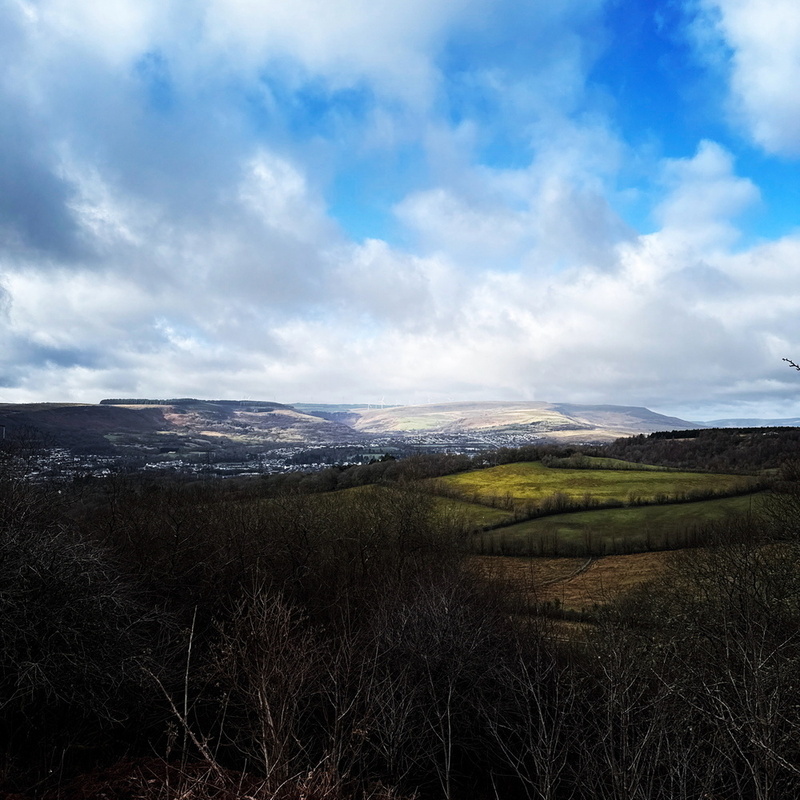 A scenic view of a valley with green fields and hills under a cloudy sky, illuminated by sunlight breaking through.